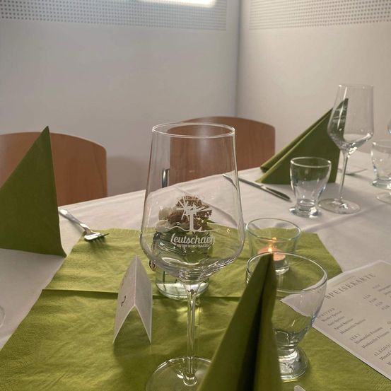 Ein Tisch mit grünen Servietten, einem Weinglas und Besteck. Auf dem Tisch befinden sich weitere Weingläser und eine Kerze. Im Hintergrund sind Stühle und eine Speisekarte.