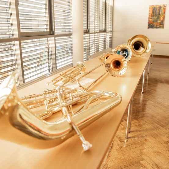 Eine Reihe glänzender Blechblasinstrumente auf einem langen Tisch in einem Musikraum. Die Instrumente sind in verschiedenen Farben.