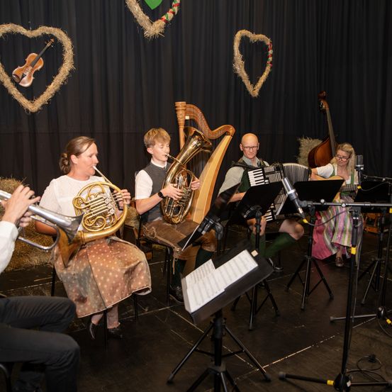 Eine Band spielt auf der Bühne, bestehend aus einem Geiger, einem Harfenisten und einem Cellisten. Der Geiger spielt mit einem Bogen, während der Harfenist und der Cellist auf Stühlen sitzen. Eine Frau sitzt auf dem Boden und spielt Akkordeon.