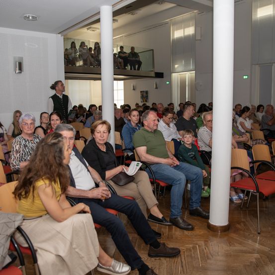 Ein Publikum in einem Theater oder Konferenzraum. Menschen unterschiedlichen Alters und Geschlechts sitzen auf Stühlen, einige stehen oder lehnen sich an Wänden. Ein Mann in einem grünen Shirt hält ein Papier, und ein Junge sitzt auf einem Stuhl vor ihnen.