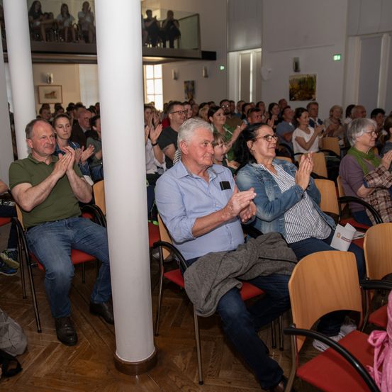 Das Publikum in einem Saal klatscht und lächelt. Ein älterer Mann in einem grünen Shirt und ein älterer Mann in einem blauen Hemd klatschen. Eine Frau in einer Jeansjacke lächelt.