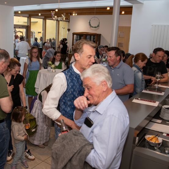 In einem überfüllten Restaurant unterhalten sich zwei Männer. Einer hält eine Flasche. Die anderen essen und unterhalten sich. Der Raum ist gut beleuchtet und mit verschiedenen Gegenständen dekoriert.