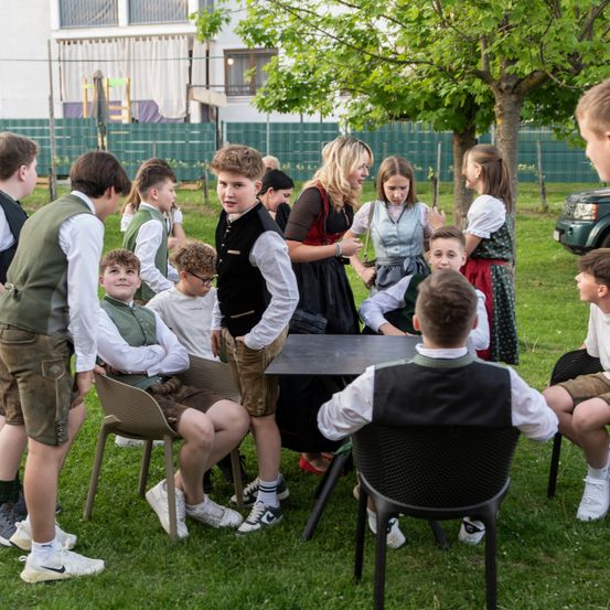 Eine Gruppe von Kindern in traditioneller Kleidung sitzt um einen Tisch in einem Grasbereich, mit einem Gebäude und einem Baum im Hintergrund.