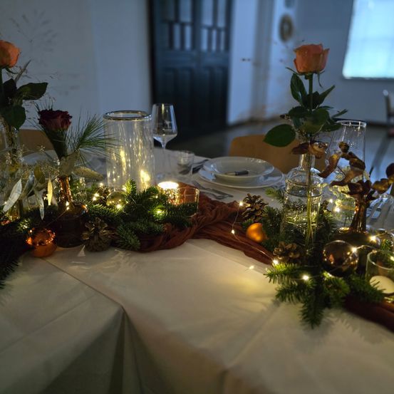 Eine festliche Tischdekoration mit Rosen, Kerzen und Weihnachtsdekoration. Der Tisch ist mit weißem Stoff bedeckt, und die Tischmitte hat eine Glasvase mit einer Rose.