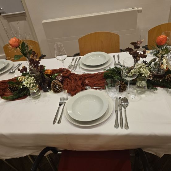 Ein Tisch für eine Dinnerparty gedeckt mit weißen Tellern, Besteck, Weingläsern und dekorativen Blumen und Tannenzapfen auf dem Tisch.