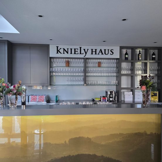 Eine Bartheke mit 'KnieLy HAUS'-Schild darüber. Regale mit Weingläsern, Flaschen und einem Waschbecken mit Wasserhahn. Zwei Vasen mit Blumen auf der Theke. Ein verschwommener Hintergrund mit Landschaft.