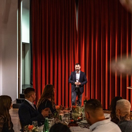 Ein Mann steht vor einem roten Vorhang und spricht ein sitzendes Publikum an. Die Umgebung deutet auf eine formelle Veranstaltung mit Blumengestecken und formeller Kleidung hin.