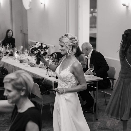 Ein schwarz-weißes Foto einer Hochzeitsfeier mit einer Braut, die Blumen hält, einem Mann im Anzug, der sitzt, und einer Frau in einem Kleid, die in der Nähe steht.