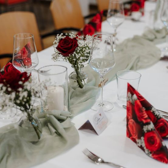 Ein Tisch ist für eine besondere Veranstaltung gedeckt. Rote Rosen und weiße Blumen sind in Vasen. Jedes Gedeck hat ein Weinglas, ein Wasserglas, ein Tuch und eine Kerze.
