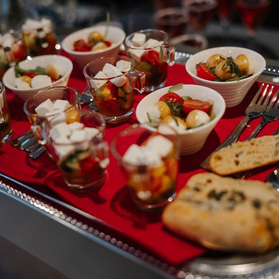 Ein Tisch mit rotem Tuch hat mehrere Glasbecher gefüllt mit einer bunten Fruchtsalat und Mozzarella-Kugeln. Eine Gabel und Brot sind auf der Seite.