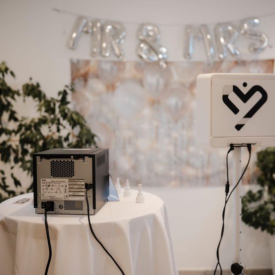 Ein Computerturm und eine Fotokabine stehen auf einem Tisch. Eine 'MR & MRS'-Ballondekoration befindet sich an der Wand dahinter.
