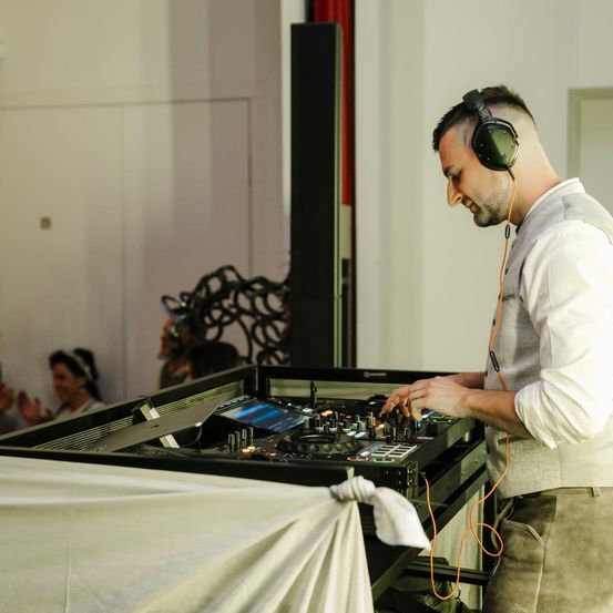 Ein Mann mit Kopfhörern und Weste bedient eine DJ-Mixkonsole bei einer Veranstaltung. Mehrere Personen sind im Hintergrund.