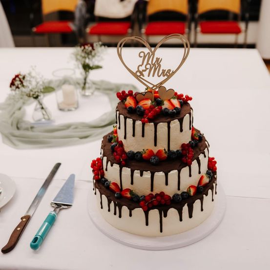 Ein dreistöckiger Hochzeitskuchen mit Schokoladen- und Beeren-Trüffeln, gekrönt von einem herzförmigen Schild, das Mr. & Mrs. sagt. Auf einem weißen Tisch mit Blumen und Stühlen im Hintergrund.