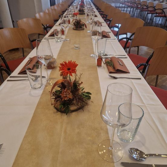 Ein langer Tisch mit Holzstühlen, jeder mit Weingläsern, Blumenarrangement und Besteck. Der Tisch ist mit einer weißen Tischdecke und einem beigefarbenen Tischläufer bedeckt.