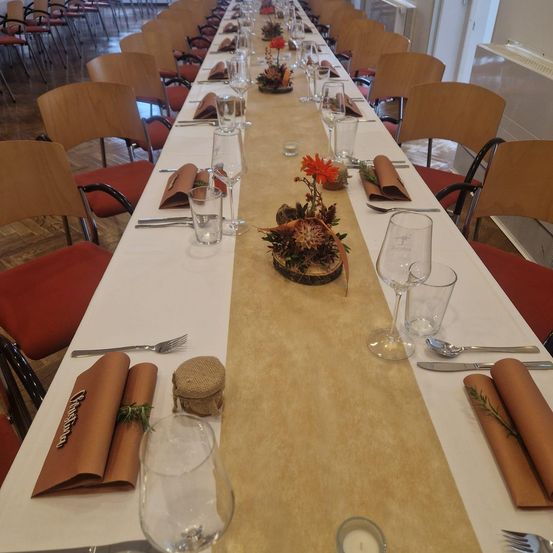 Ein langer Esstisch mit gedeckten Plätzen, einschließlich Weingläser, Besteck und Servietten. Der Tisch ist mit einer zentralen Blumendekoration und einem Tischläufer geschmückt. Die Stühle sind in Reihen auf beiden Seiten angeordnet.