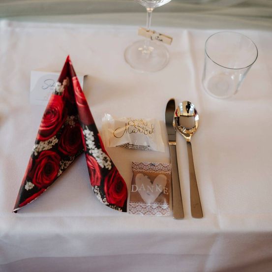 Eine Tischdekoration umfasst ein rotes Blumenserviette, ein Glas, Besteck und einen klaren Glasbecher. Ein Namensschild ist auf dem Tisch platziert.