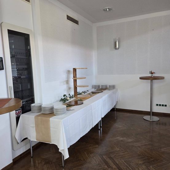 Ein Raum mit einem langen Tisch, der für eine Mahlzeit gedeckt ist, mit Tellern, einer weißen Tischdecke, einer hölzernen Wand und einem runden Tisch auf der rechten Seite.