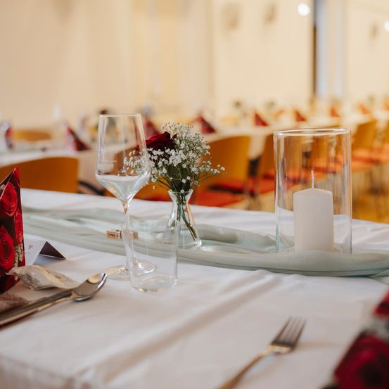 Ein gedeckter Tisch mit weißer Tischdecke, einem Weinglas, einer Kerze und einer Blumenarrangement. Das Ambiente ist elegant mit anderen Tischen und Stühlen im Hintergrund.
