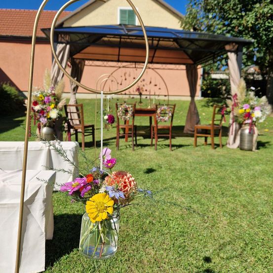 Outdoor-Hochzeitsaufbau mit bunten Blumen, einem hölzernen Pavillon und Tischen unter einem Vordach. Ein großer goldener Ring hängt im Vordergrund.