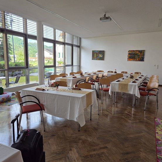 Ein Esssaal mit Holzboden und großen Fenstern, Tische für eine Mahlzeit gedeckt, Stühle darum herum und zwei Gemälde an der Wand.