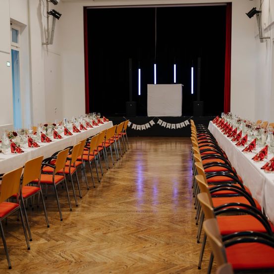 Ein Bankettsaal mit Bühne und Tischen, die für eine große Veranstaltung vorbereitet sind. Lange Tische mit weißen Tischdecken, roten Servietten und Gläsern. Stühle sind in Reihen angeordnet. Eine schwarze Kulisse und rote Vorhänge rahmen die Bühne ein.