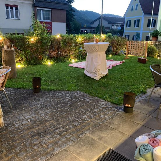 Ein abendlicher Aufbau in einem Garten, mit einem in Weiß bedeckten Tisch, umgeben von Stühlen und einer Decke. Zwei Laternen beleuchten den Bereich.