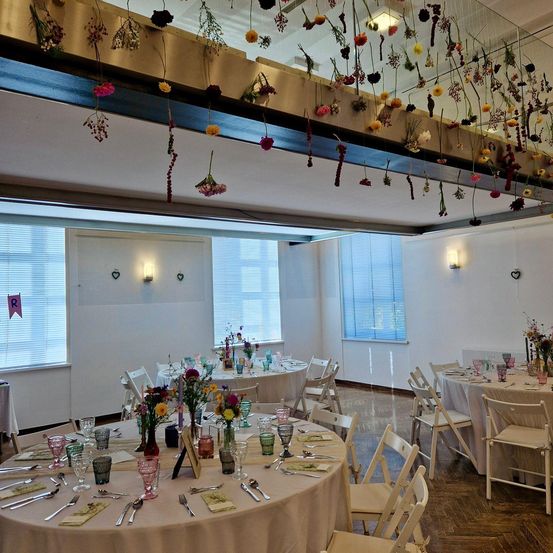 Ein Indoor-Essbereich mit mehreren Tischen, die mit Blumen und Besteck gedeckt sind. Eine Glasdecke hält einen Strauß getrockneter Blumen. Der Raum hat Fenster mit Jalousien und eine Wand mit Herz-Design.