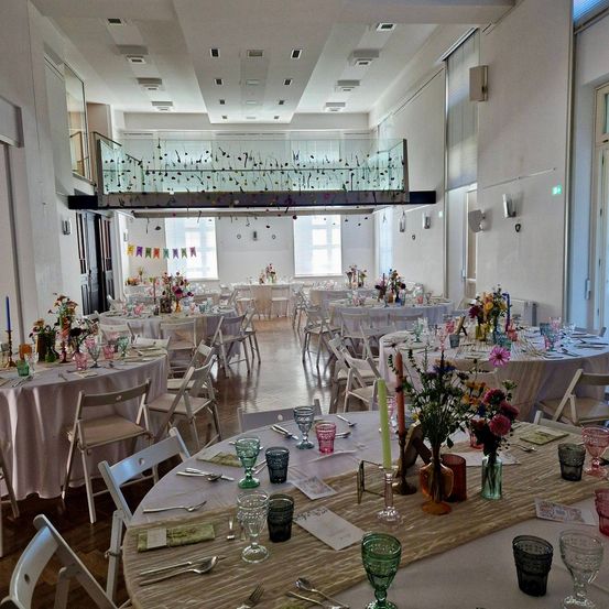 Ein Bankettsaal ist mit mehreren runden Tischen eingerichtet, die jeweils mit Blumengestecken, Gläsern und Besteck geschmückt sind. Der Raum hat hohe Decken, weiße Wände und eine Galerie oben.