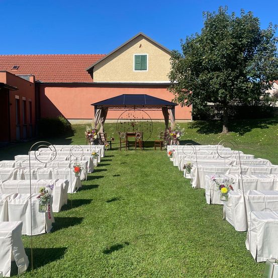 Eine Hochzeitszeremonie ist auf einer Wiese mit weißen Stühlen, Blumenarrangements und einem Pavillon eingerichtet. Ein Gebäude mit rotem Dach und grünen Fensterläden steht dahinter.