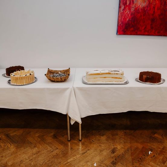 Zwei Tische mit weißen Tischdecken sind mit verschiedenen Kuchen gedeckt. Jeder Tisch hat eine Vielfalt an Desserts. Ein abstraktes Gemälde hängt an der Wand.