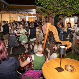 Eine Gruppe von Menschen in traditioneller bayerischer Kleidung tanzt und spielt Musikinstrumente in einem Garten. Eine Harfe und ein Akkordeon sind vorhanden, mit einer Lampe und Tischen in der Nähe.