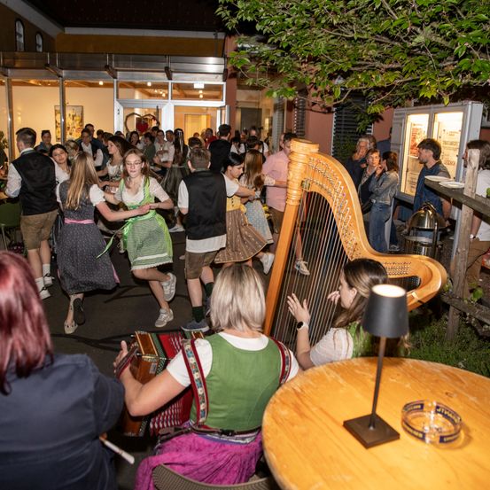 Eine Gruppe von Menschen in traditioneller bayerischer Kleidung tanzt und spielt Musikinstrumente in einem Garten. Eine Harfe und ein Akkordeon sind vorhanden, mit einer Lampe und Tischen in der Nähe.