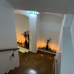 An interior view of a staircase with two wooden shelves, each adorned with candles and a vase, illuminated by warm light. A green exit sign is visible above.