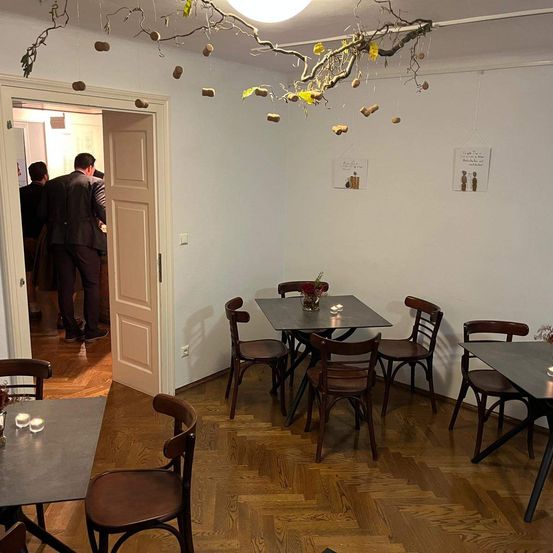 Ein leeres Restaurant mit Holzböden und weißen Wänden. Es gibt Holzstühle und Tische mit Kerzen. Ein Zweig mit Blättern hängt von der Decke.