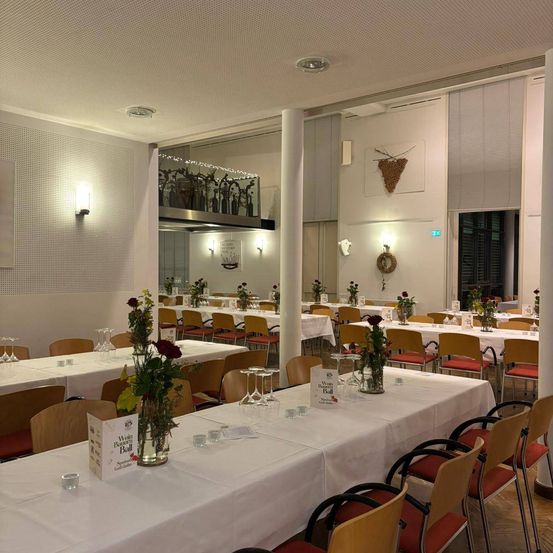 Ein Esszimmer ist für eine besondere Veranstaltung dekoriert. Auf den Tischen sind weiße Tischdecken, Blumen und Gläser arrangiert. Der Raum hat Holzstühle, eine Säule und dekorative Gegenstände an den Wänden.