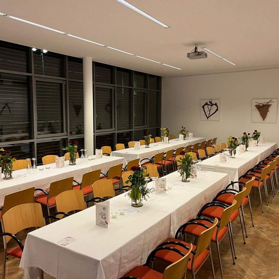 Ein Konferenzraum ist mit mehreren langen Tischen ausgestattet, die mit weißen Tischdecken bedeckt sind, jeder mit Blumen und Stühlen darum herum. Der Raum hat große Fenster mit Jalousien, und zwei gerahmte Kunstwerke hängen an der Wand. Der Raum ist gut beleuchtet durch Deckenleuchten.
