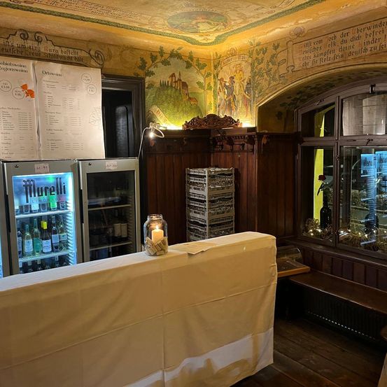 Ein Raum mit Holzwänden und einem abgedeckten Tisch hat zwei Glaskühlschränke gefüllt mit Getränken. Ein Menübrett hängt an der Wand und eine Kerze brennt in einem Glastopf.