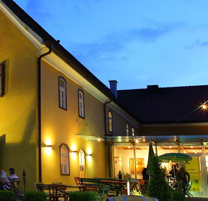 Ein gelbes Gebäude mit mehreren Fenstern ist bei Dämmerung beleuchtet. Menschen sitzen draußen auf Stühlen. Es gibt einen Fass im Vordergrund.