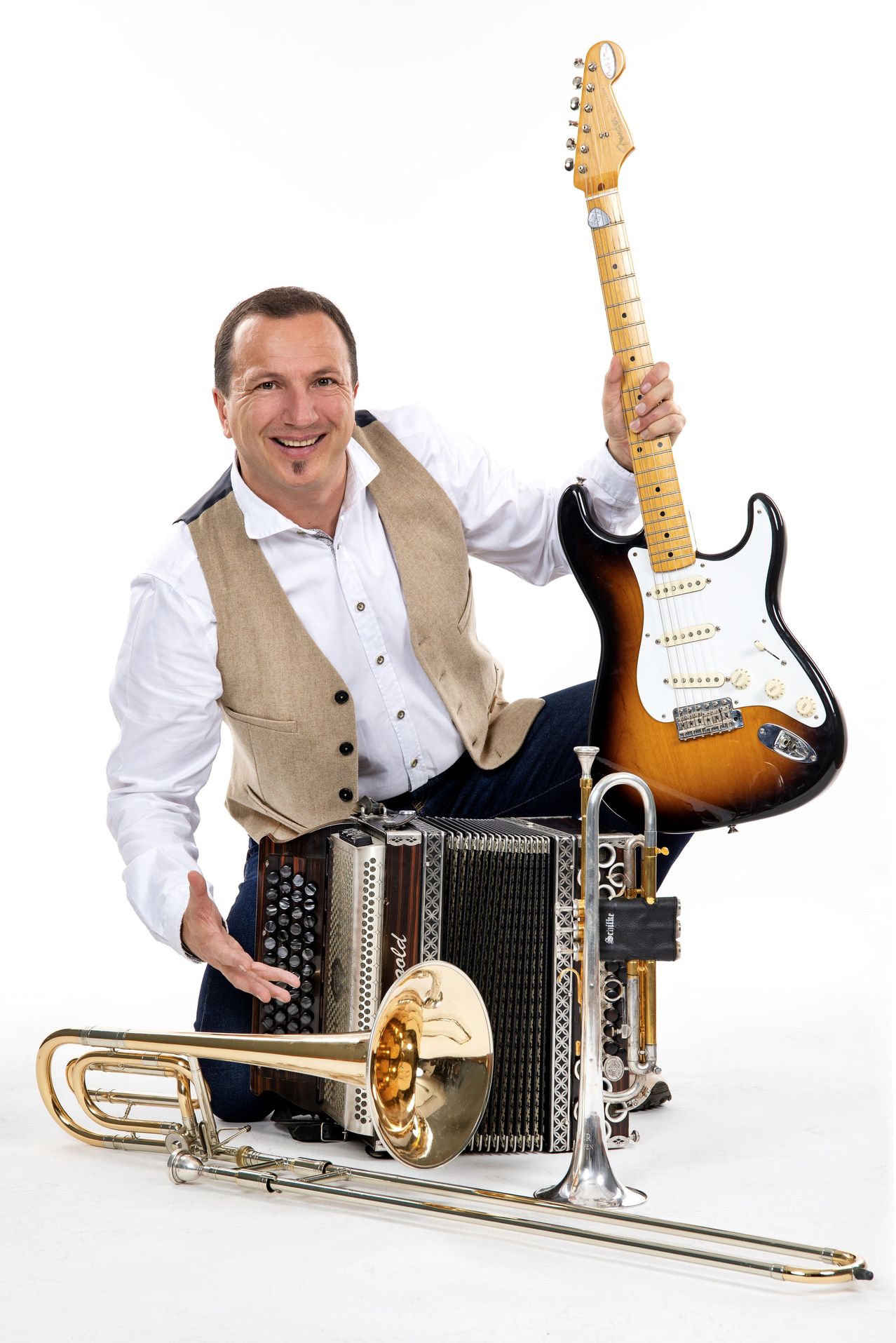 Ein Mann mit einer Gitarre, einem Akkordeon und einer Trompete, lächelt und posiert für ein Foto.