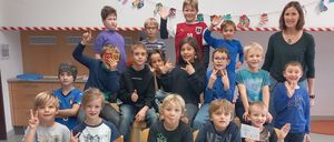 Ein Klassenfoto von Schülern, die verschiedene Gesten machen. Eine Lehrerin steht im Hintergrund und lächelt. Die Schüler tragen Freizeitkleidung und sitzen auf Stühlen.