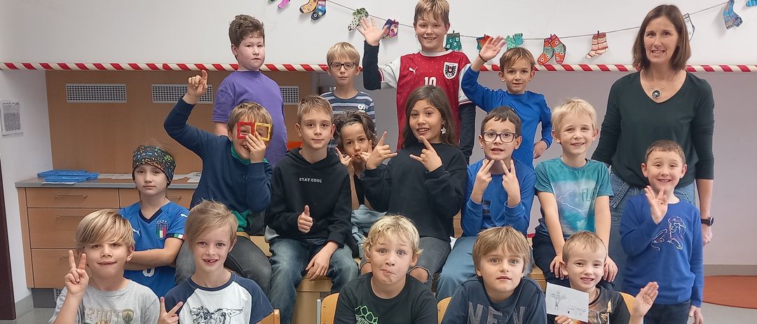 Ein Klassenfoto von Schülern, die verschiedene Gesten machen. Eine Lehrerin steht im Hintergrund und lächelt. Die Schüler tragen Freizeitkleidung und sitzen auf Stühlen.