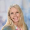 Eine Frau mit langen blonden Haaren lächelt und posiert für ein Foto. Sie trägt ein grünes Oberteil und ein rosa Blumenhemd. Ihre Haare sind leicht gewellt und sie hat einen Muttermal auf ihrer rechten Wange. Der Hintergrund ist unscharf.