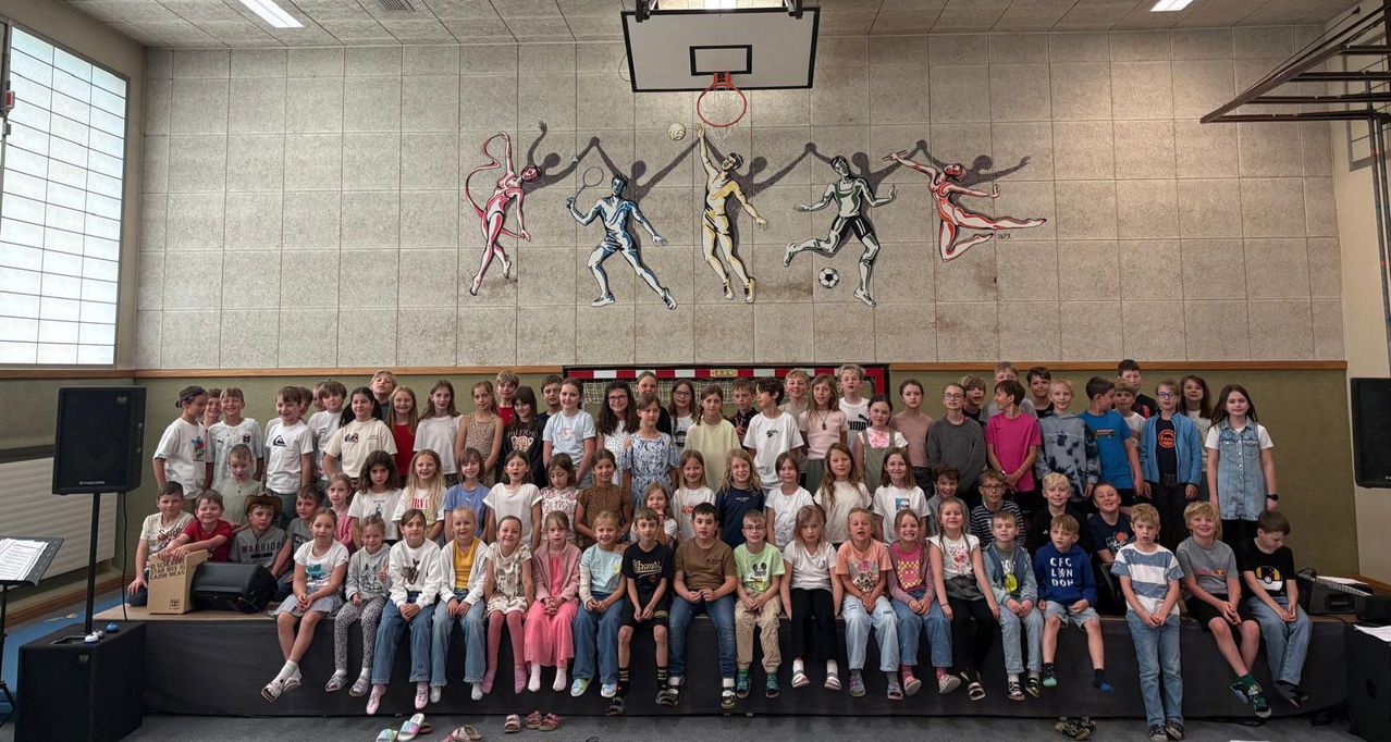 Eine große Gruppe von Kindern und Jugendlichen posiert für ein Foto in einer Turnhalle mit Sportmurals an der Wand.