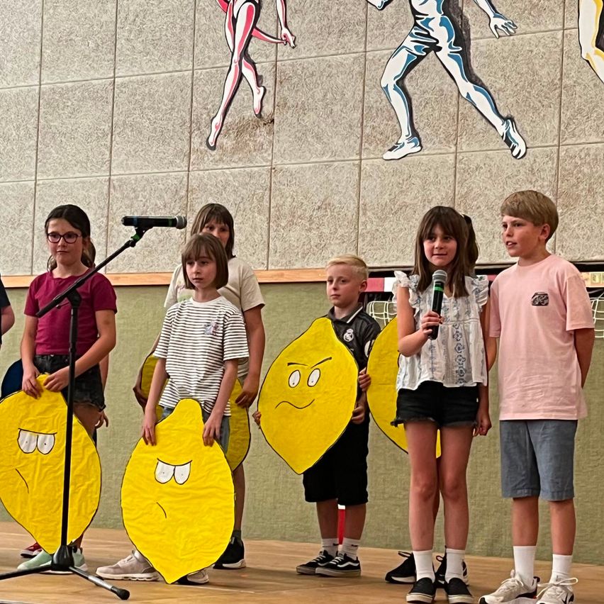 Mehrere Kinder stehen auf einer Bühne und halten Papierausschnitte in Form von Zitronen. Ein Mädchen hält ein Mikrofon. Sie stehen vor einer Wand mit bunten Illustrationen.