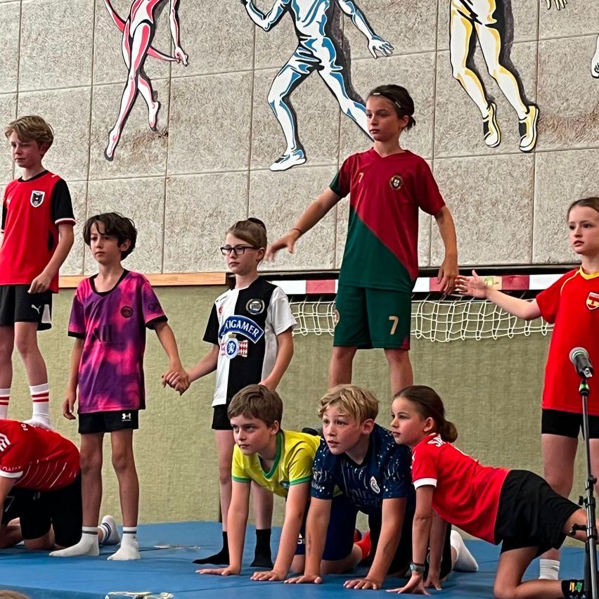 Mehrere junge Kinder in Sportuniformen hocken auf einem blauen Matten, möglicherweise in einer Turnhalle. Ein Mädchen in einem roten Trikot hält ein Mikrofon. An der Wand dahinter befinden sich Wandgemälde von rennenden Figuren.