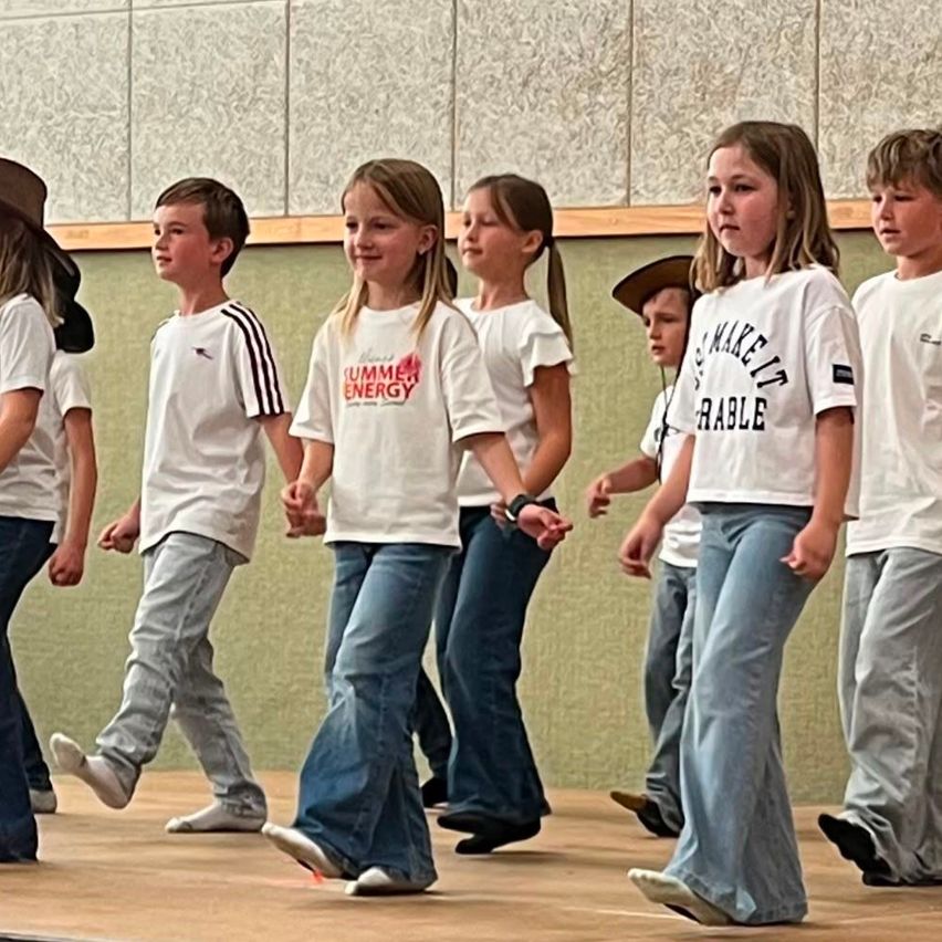 Eine Gruppe von Kindern in weißen T-Shirts und Jeans läuft auf einer Bühne, einige halten sich an den Händen. Das Mädchen in der Mitte hat eine Uhr am Handgelenk.