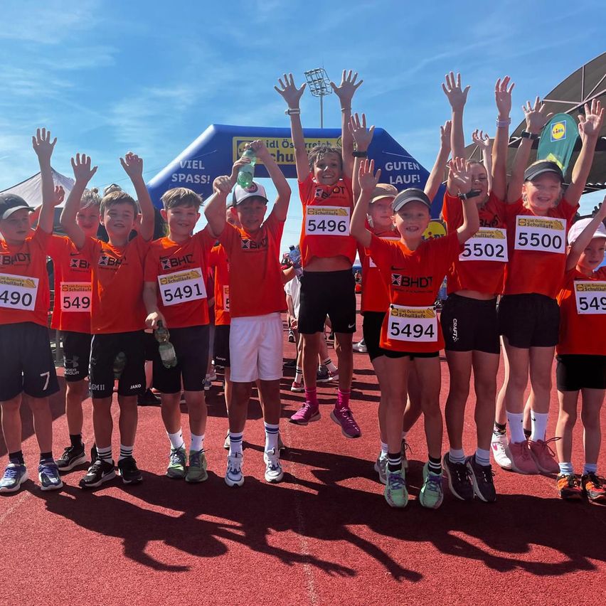 Eine Gruppe von Kindern in orangefarbenen T-Shirts mit Nummern posiert für ein Foto auf der Laufbahn. Sie tragen Laufschuhe und Mützen, einer hält eine Flasche. Der Himmel ist blau.