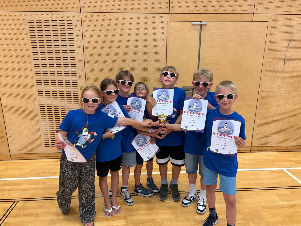 Eine Gruppe von Kindern, die Urkunden und einen Pokal in einer Turnhalle halten. Sie tragen passende blaue T-Shirts, Sonnenbrillen und Turnschuhe.