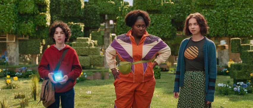 Drei Personen stehen auf einer Wiese mit einer grün beschnittenen Hecke im Hintergrund. Die Person in der Mitte trägt ein orangefarbenes Jumpsuit, während die anderen lässige Kleidung tragen.