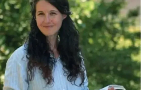 Eine Frau mit lockigem Haar lächelt und hält ein offenes Buch in der Hand. Der Hintergrund ist unscharf mit grünen Bäumen.